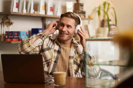 Lifestyle portrait of happy, excited young man, sitting in coffee shop with laptop and headphones, dancing on his chair and enjoying great quality sound, streaming favourite songs.