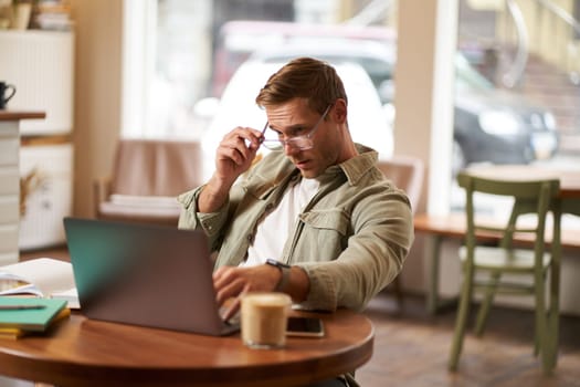 Portrait of a man working in cafe, looking surprised at camera, taking off glasses to stare puzzled at laptop screen, has a question about something on his computer. concept of remote jobs.