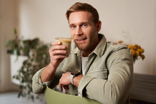 Portrait of good-looking young man with cup of coffee, sitting on chair in cafe, smiling and relaxing with his cappuccino drink.