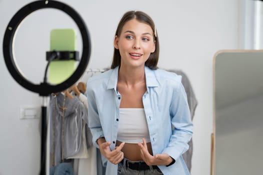 Young woman making fashion video content for social media. Blogger smiles to camera using selfie stick and light ring while making persuasive online clothing sell to audience or followers. Unveiling