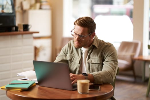 Portrait of businessman in glasses, sits in cafe with laptop, looks concentrated at screen with his project task, works remotely from coffee shop.