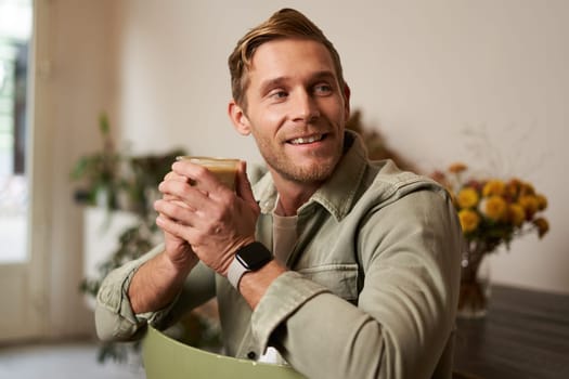 Portrait of good-looking young man with cup of coffee, sitting on chair in cafe, smiling and relaxing with his cappuccino drink.