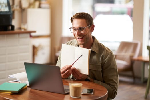 Portrait of young handsome man in glasses, private tutor teaching student online, pointing at his notebook, showing something, video chats via laptop application, working remotely in cafe.