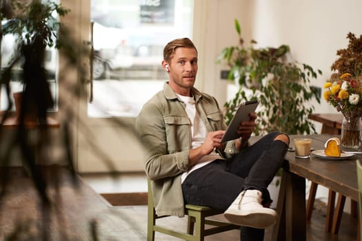 Portrait of young man sitting in cafe, spending time alone in coffee shop, reading on digital tablet, watching video, listening music in wireless headphones. Lifestyle and people concept.