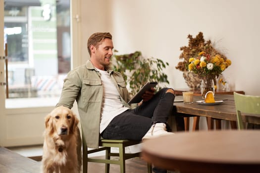 Portrait of handsome young man sits in cafe with his dog, petting golden retriever, reading news on tablet, relaxing with cup of coffee.