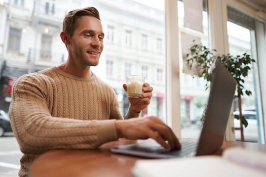 Portrait of young male businessman in cafe, drinking coffee and using his laptop, working from co-working space, sitting near the window and looking at computer screen.