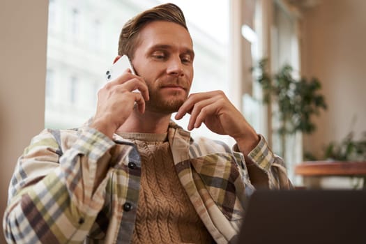 Image of young professional, cafe visitor working, sitting with laptop and talking on mobile phone to client. A guy making an enquiry buying something online, calling a friend.