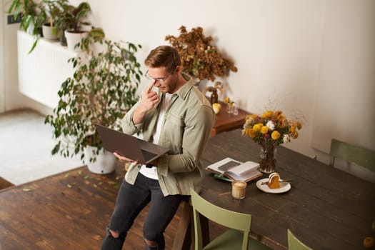 Image of young man, successful businessman sitting on table, looking at laptop, working in an office, looking concentrated on the project, doing job task, drinking coffee.