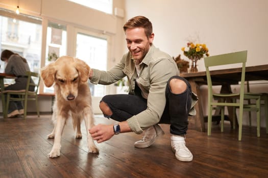 Portrait of happy man spending time with his dog in pet-friendly cafe, playing and petting golden retriever, having fun, giving him a treat.