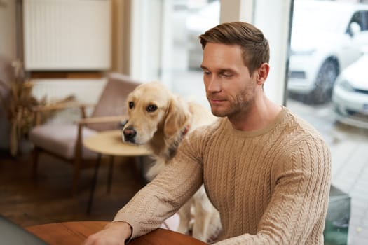 Handsome young man, digital nomad sitting with his dog in a pet-friendly cafe, working on laptop while a golden retriever watches him.