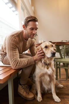 Close up portrait of handsome man with his dog, sitting in cafe. A guy drinks his coffee and touches golden retriever.
