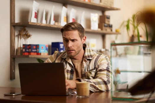 Image of young business owner, man sitting in cafe with laptop, looking confused at screen, digital nomad trying to solve the task, programmer working from coffee shop.