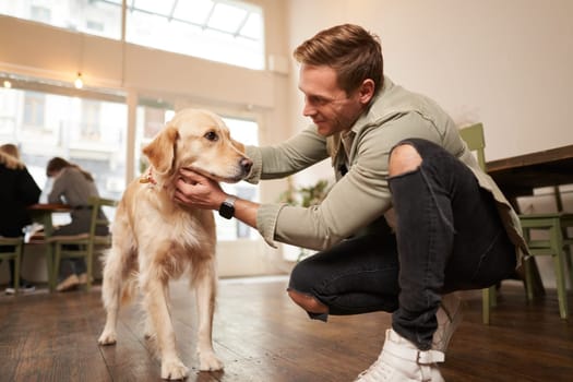 Close up portrait of handsome happy man petting his cute dog in a pet-friendly cafe. Coffee shop visitor with his golden retriever.