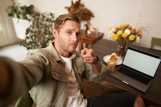 Confident, handsome man, pointing finger at his mobile phone, taking selfie, frowning sassy, sitting in cafe with laptop, drinking coffee.