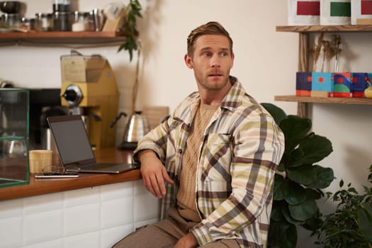 Small business concept. Handsome businessman, cafe owner, sitting near counter with laptop and smartphone, working in coffee shop.