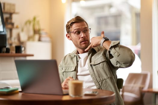 Portrait of young handsome man in glasses, businessman working in cafe, sitting with laptop, showing phone call hand gesture, advertising his service.