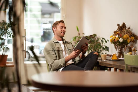 Portrait of handsome smiling young man, sitting in coffee shop, using digital tablet, video chats with someone from a cafe, wearing wireless headphones.