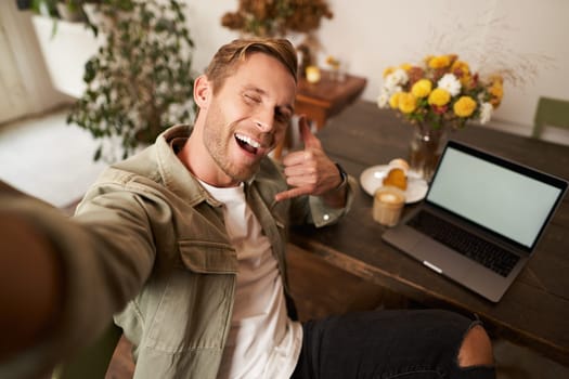 Handsome stylish young man takes selfie in cafe while working or studying remotely with laptop, shows call phone hand sign and smiling at mobile phone camera.