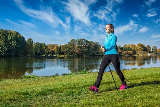 Nordic walking adventure and exercising - young woman hiking with nordic walking poles in park along river
