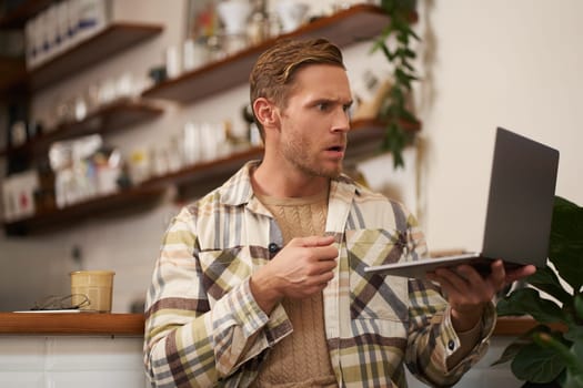 Portrait of man looking stressed and concerned at laptop screen, clench fist and frowns with angry face expression, sits in cafe with cup of coffee.