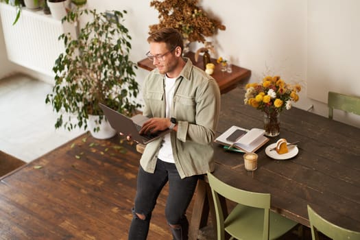 Handsome manager, man in glasses sits on table, holds laptop and types, sends message, works on project, drinks coffee, spends time at co-working space or an office.