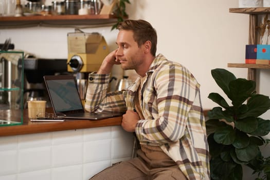 Image of adult man looking complicated at laptop screen, staring puzzled at his monitor, doing difficult task online, sitting in cafe, drinking coffee and working.