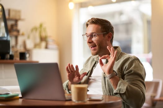 Portrait of young man, teacher, online tutor in glasses, showing okay, ok hand sign, giving lessons remotely from cafe, sitting with laptop, talking to someone on video chat.