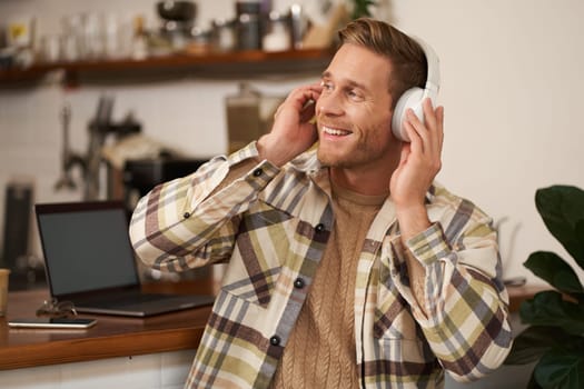 Portrait of handsome happy guy, freelancer working and listening to music in wireless headphones, dancing on his chair in cafe, enjoying favourite song in earphones.