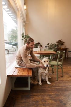 Handsome man sitting in cafe with his dog. A guy drinks coffee and strokes golden retriever. Animal-friendly coffeeshop.