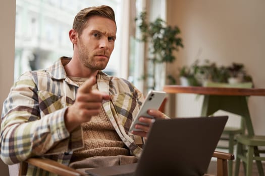 Portrait of man with serious face, sitting in cafe with laptop and smartphone, pointing at you with warning, scolding you, frowning with disapproval.