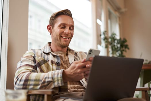 Close up portrait of handsome smiling man with laptop, cafe visitor sitting in chair, using smartphone, looking happy, messaging with friends while enjoying cup of coffee.