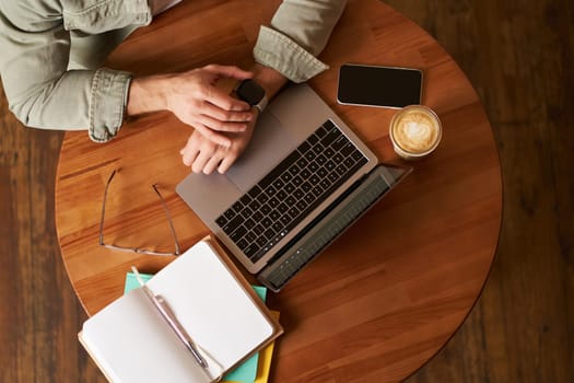 Top view of male hands, man checking time or message on his digital watch, sitting in a cafe with laptop, mobile phone and notebook, drinking coffee.