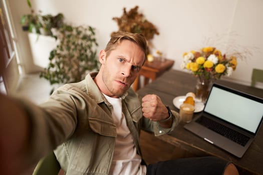 Portrait of serious man takes selfie with fist and threatening face, sits in cafe, poses in coffee shop in front of table with laptop and glass of coffee.