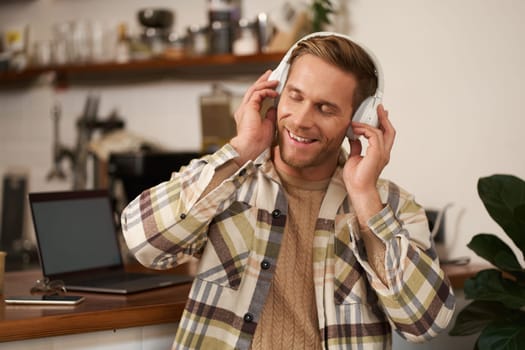 Portrait of young man sitting with laptop in cafe, listening music in headphones, smiling with eyes closed, pleased by the sound quality.