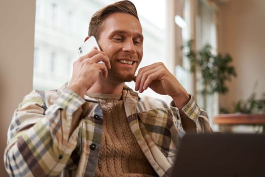 Handsome businessman in coffee shop, sitting in cafe with laptop and calling someone, Man talking on the phone with client, making enquiry about project, talking to coworker while working remotely.