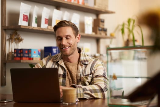 Portrait of cafe owner, young man sitting in coffee shop with laptop, working on data project, looking at screen concentrated. Lifestyle and business concept