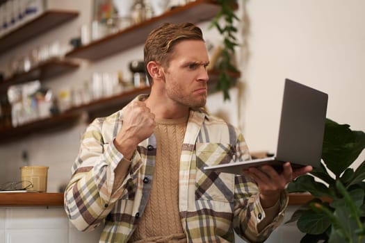 Portrait of angry man in cafe, holding laptop, staring at screen and showing clenched fist during video chat, online meeting.