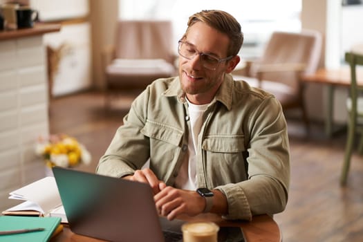 Remote work and freelance concept. Handsome man in glasses sits with notebook and laptop, studying online, working remotely from coffee shop, sitting in cafe with computer.