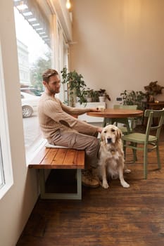Vertical shot of young man, owner of golden retriever, sitting in cafe with and stroking his dog under the table, drinking coffee in a coffeeshop.
