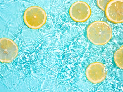 Lemon slices in clean transparent water over blue background with copy space. Water splashing on blue water surface in sunlight. Top view or flatlay. Summer, vacation, healthy eating concept