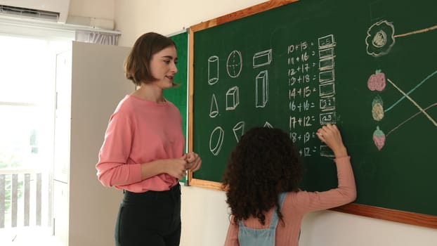 Skilled caucasian teacher looking at african girl writing answer on blackboard at elementary school. Professional student walking at in front of class to answer quiz in elementary classroom. Pedagogy.