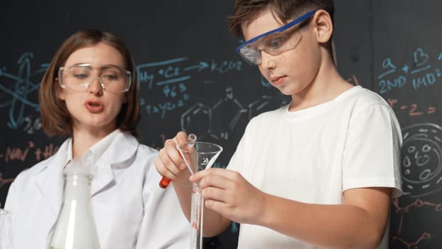 Close up of caucasian boy mixing chemical liquid while teacher giving advice. Instructor wearing lab suit looking for diverse student at table with beaker filled with colored solution. Erudition.