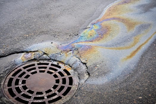 The oil slick on the asphalt road is contributing to water pollution as it drains into the storm drain