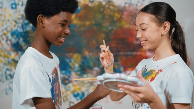 Smiling diverse children using paint brush painted color on each other white shirt shirt at colorful stained wall in art lesson. Represent exchanging experience, learning each other. Edification.