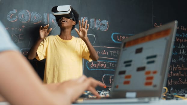 Closeup of school girl hand coding program while african student using VR interact with metaworld. Smart boy wearing virtual reality glasses and playing video games at innovative class. Edification.