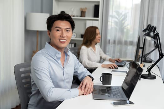 Profile smiling of smart Asian businessman looking camera to pose at modern office. Blurry background woman colleague working on pc to check email marketing plan analysis report at desk. Infobahn.