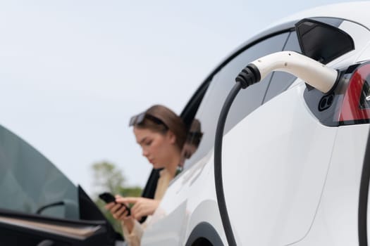 Young woman use smartphone to pay for electricity at public EV car charging station green city park. Modern environmental and sustainable urban lifestyle with EV vehicle. Expedient