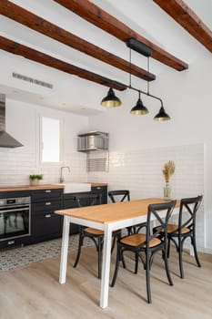 Scandinavian style kitchen with wooden long table and black chairs with kitchen island and modern built-in appliances with sink. Concept of a minimalist interior and zoning in a new house.