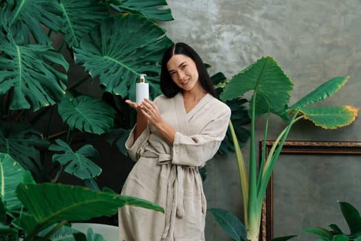 Tropical and exotic spa garden with bathtub in modern hotel or resort with woman in bathrobe holding beauty skincare product while enjoying leisure lush with greenery foliage background. Blithe