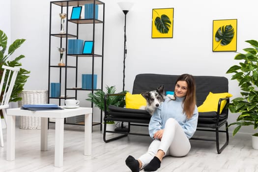 A happy teenage girl is sitting on the floor of her home in the room while the dog is lying on the sofa and her paws rest on her shoulder. Purebred Border Collie dog in shades of white and black, and long and fine hair. Living room in a modern styling.